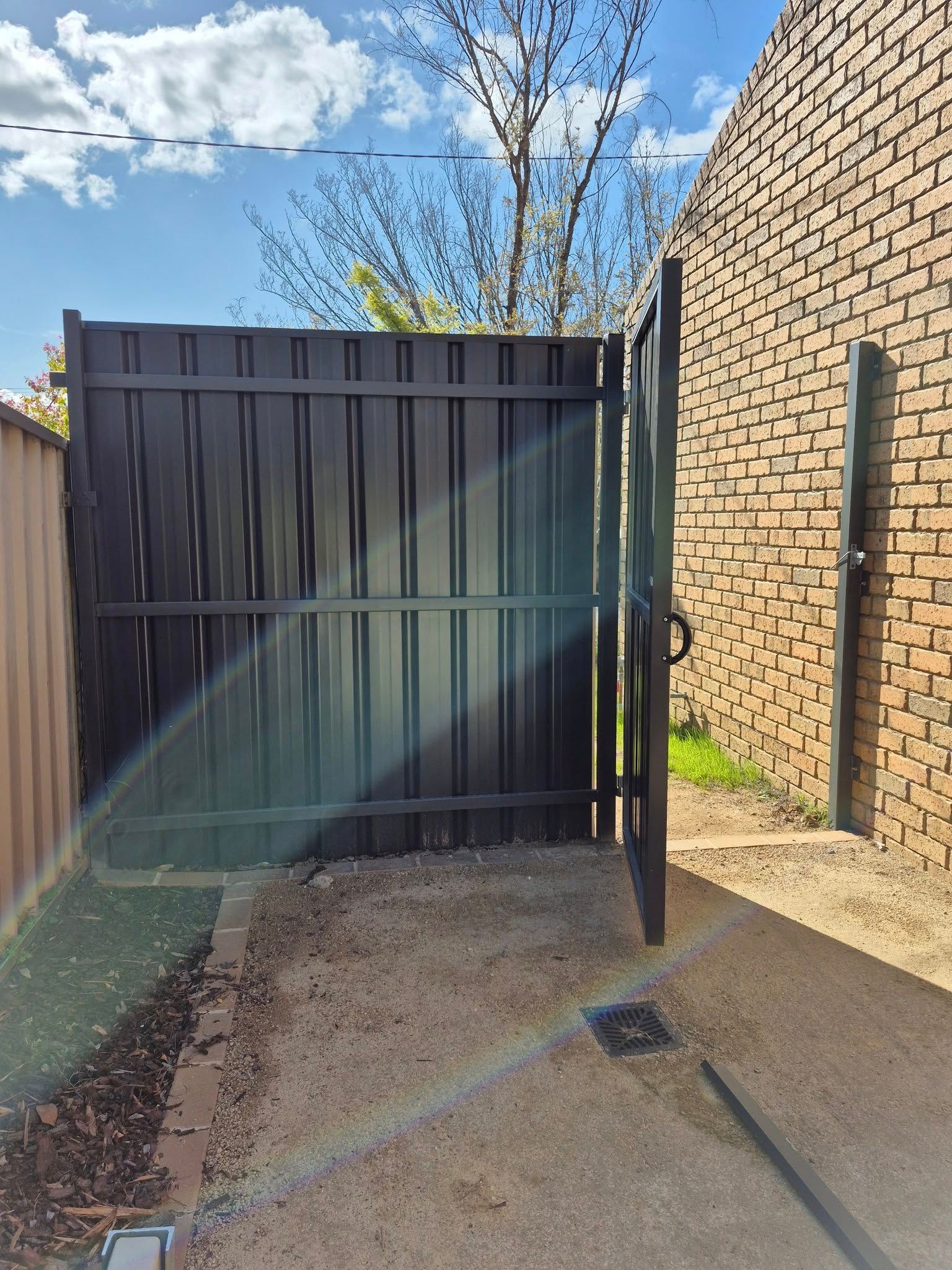 Black metal gate open, leading to gravel area, adjacent to a brick wall. Bright sunlight — Precision Fencing & Landscaping in Thurgoona, NSW