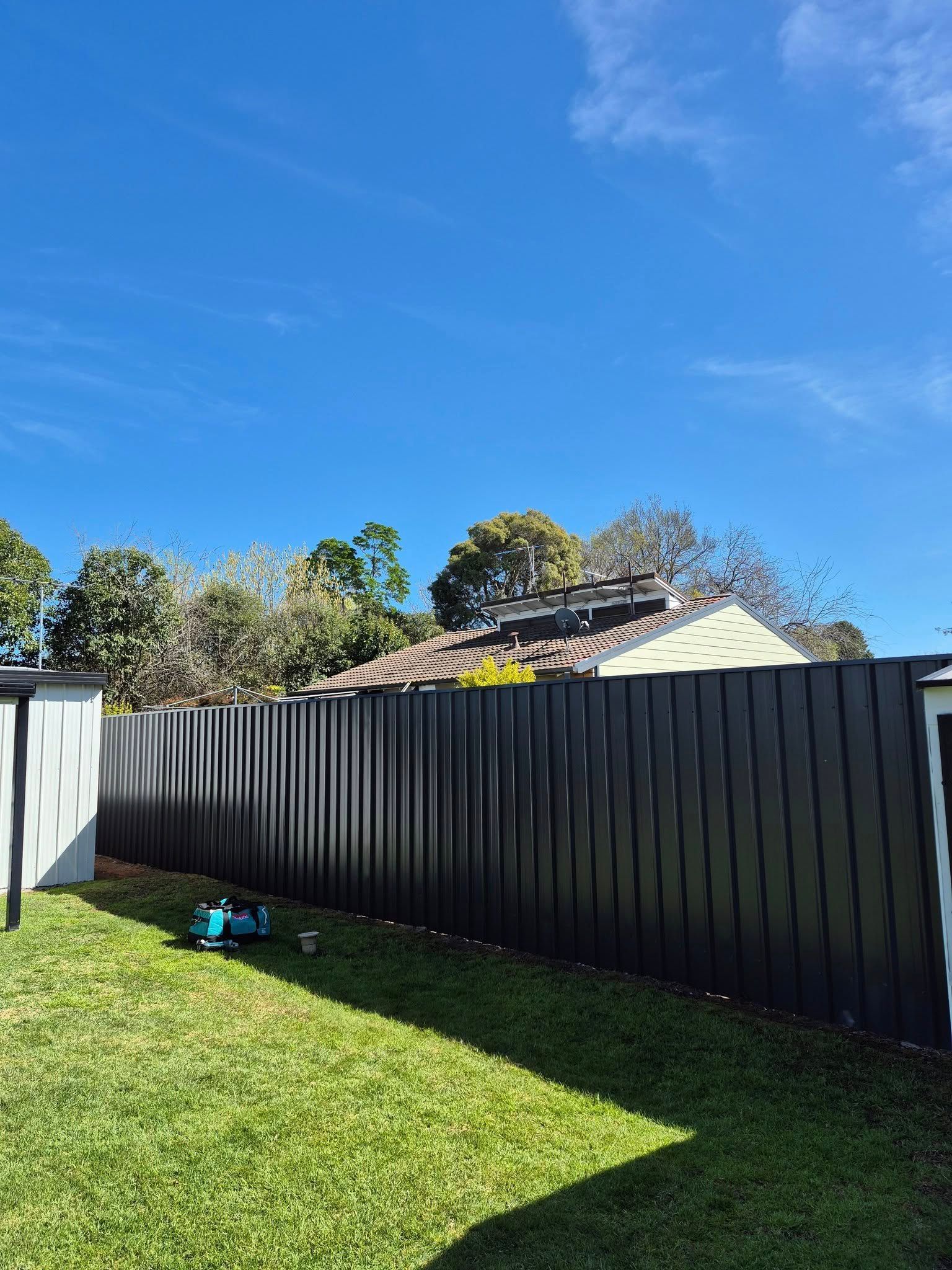 A black corrugated fence lines a backyard, blue sky overhead, with a lawnmower on the green grass — Precision Fencing & Landscaping in Thurgoona, NSW