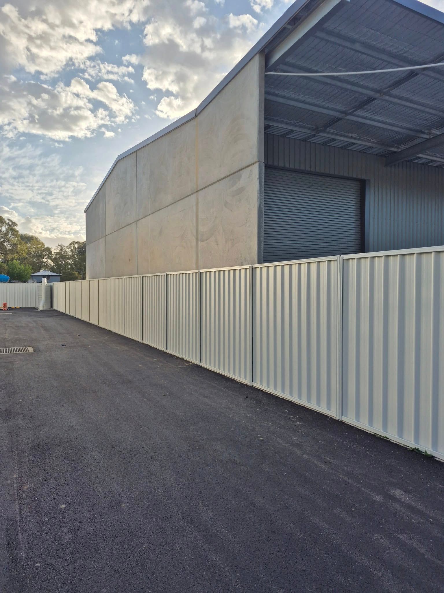 A corrugated metal fence runs along a paved driveway next to a warehouse — Precision Fencing & Landscaping in Thurgoona, NSW
