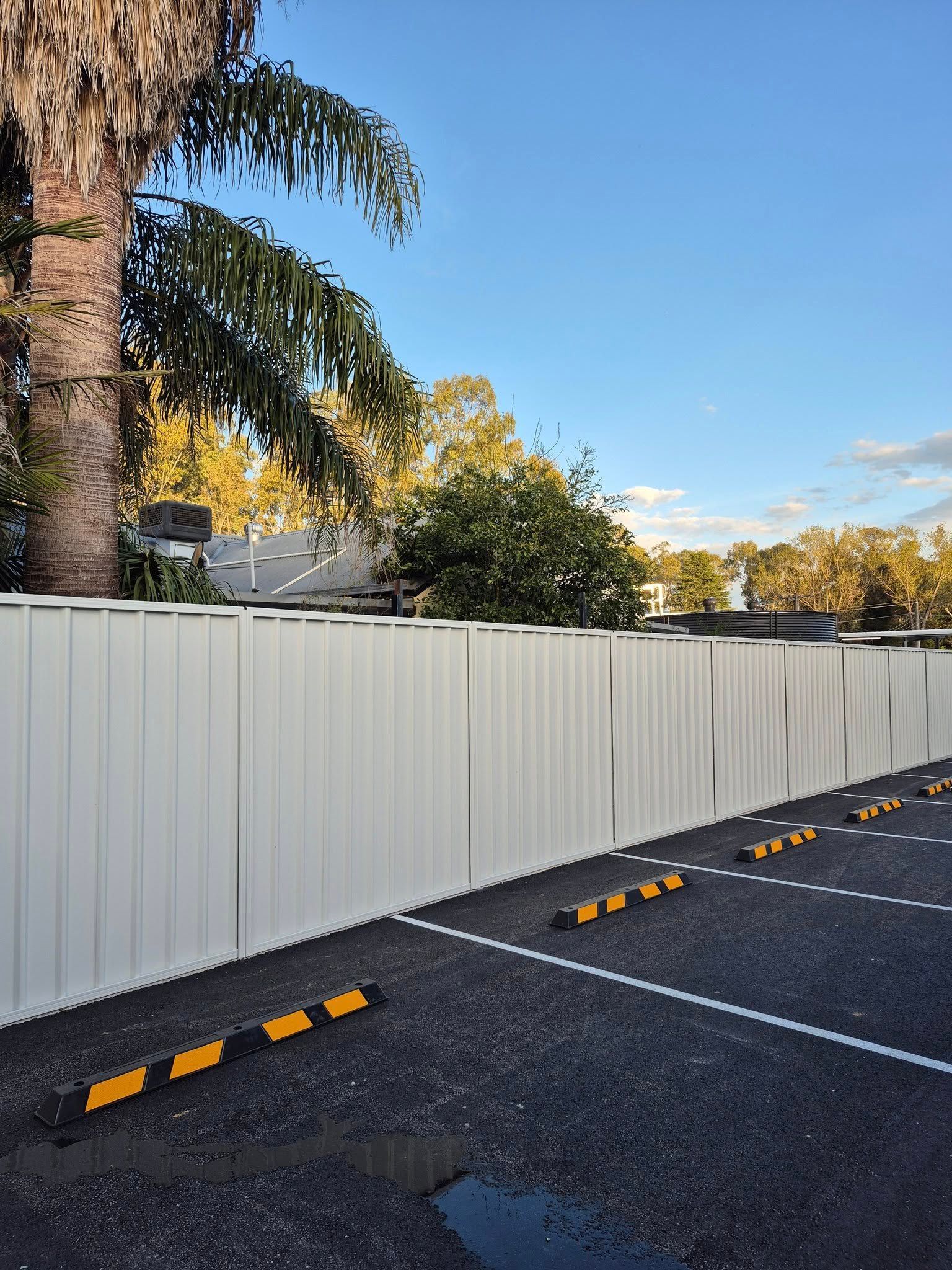 White fence bordering a parking lot with yellow and black parking stops, under a partly cloudy blue sky — Precision Fencing & Landscaping in Thurgoona, NSW