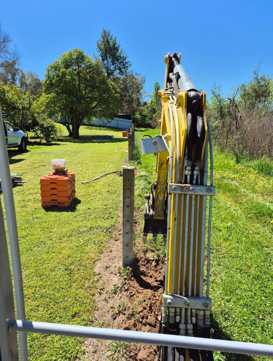 Yellow Excavator Digging a Trench Along a Fence Line in a Grassy Yard — Precision Fencing & Landscaping in Thurgoona, NSW