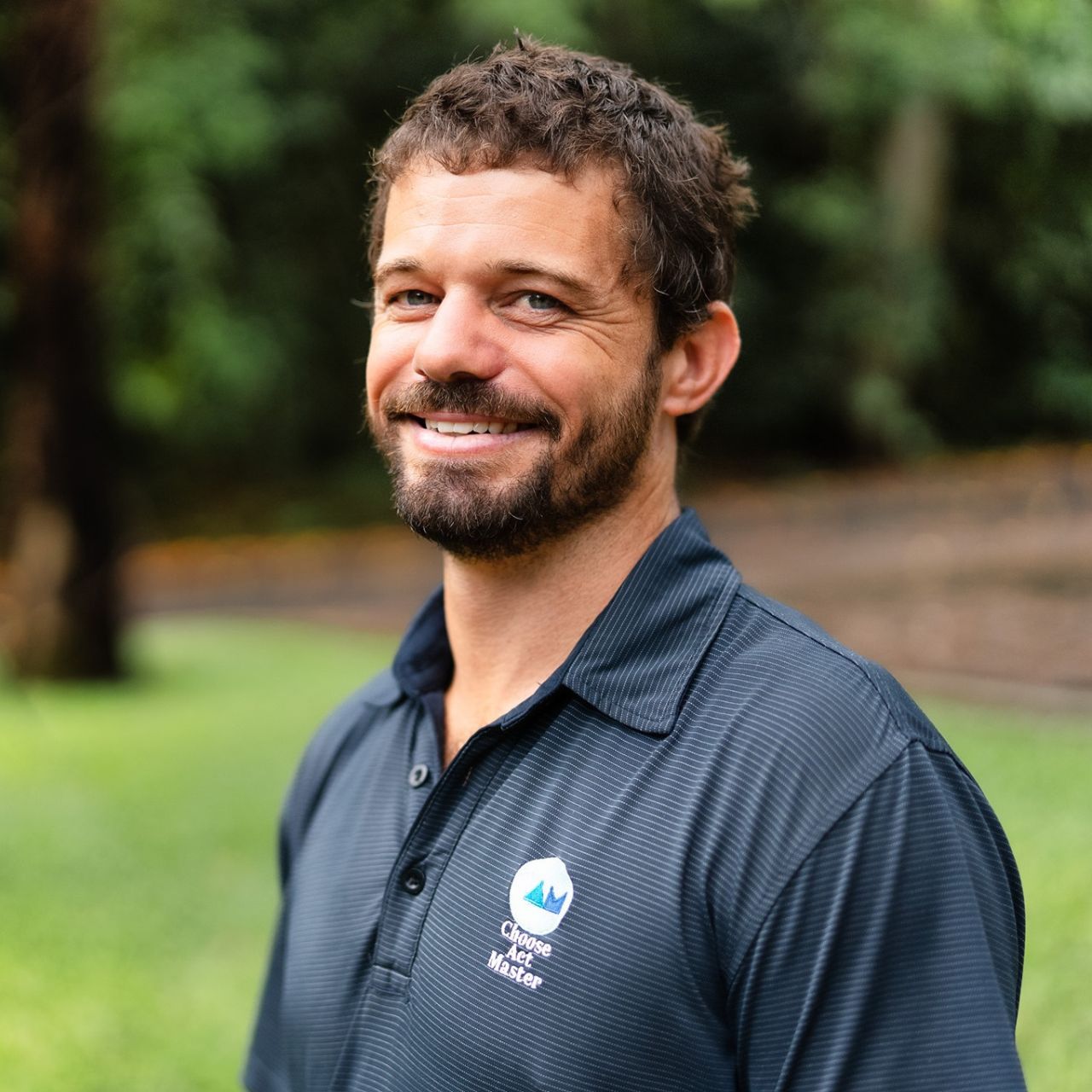Cameron Matthew from Choose Act Master Performance Coaching - a man with a beard is wearing a blue shirt and smiling .