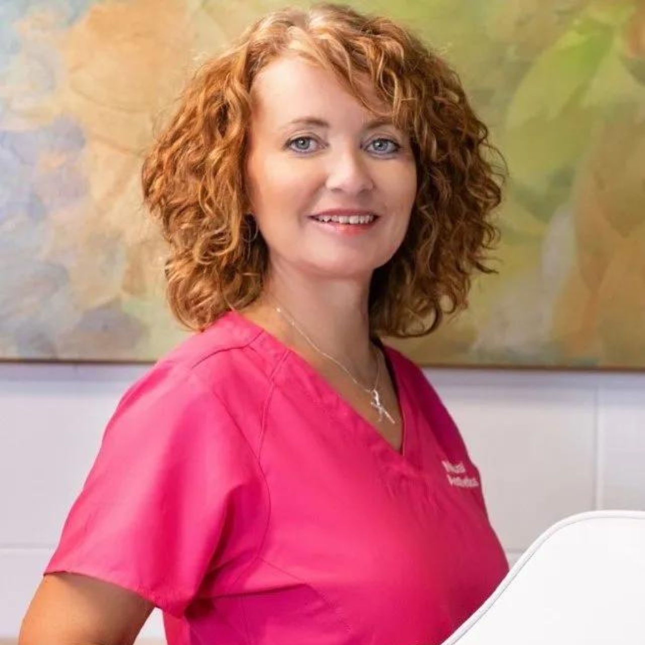 Joyce Coyle from Natural Aesthetics - a woman in a pink scrub top is standing in front of a painting .