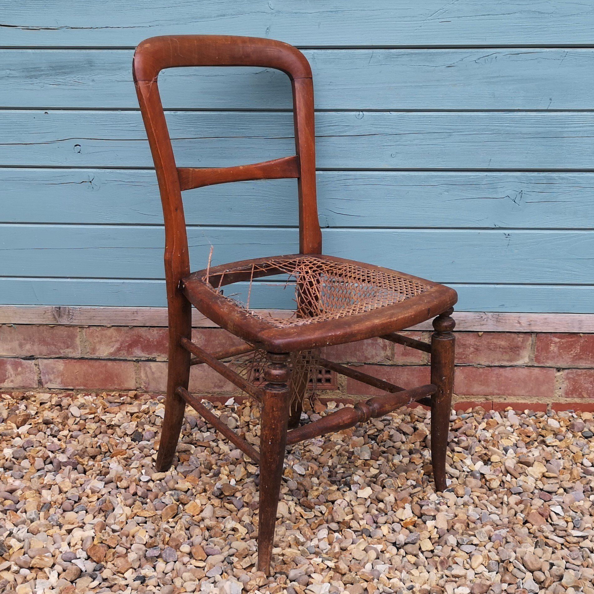 A wooden chair with a broken seat is sitting on gravel in front of a blue wall