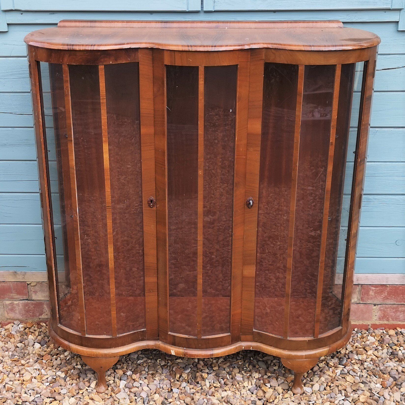 A wooden cabinet with glass doors is sitting on gravel in front of a blue building