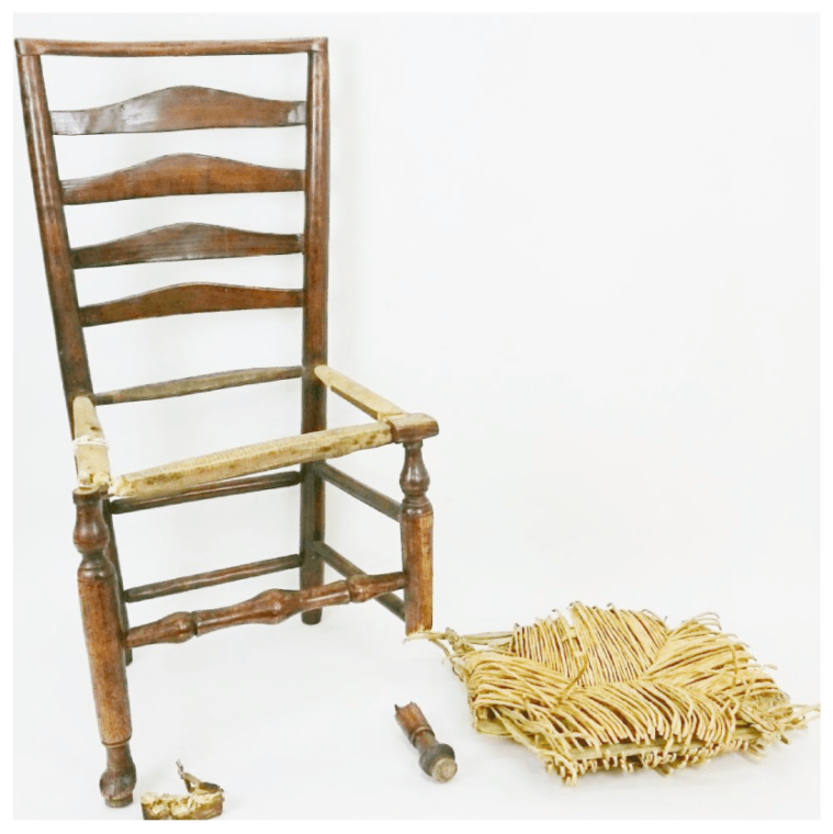 A wooden chair with a broken seat sits next to a pile of straw