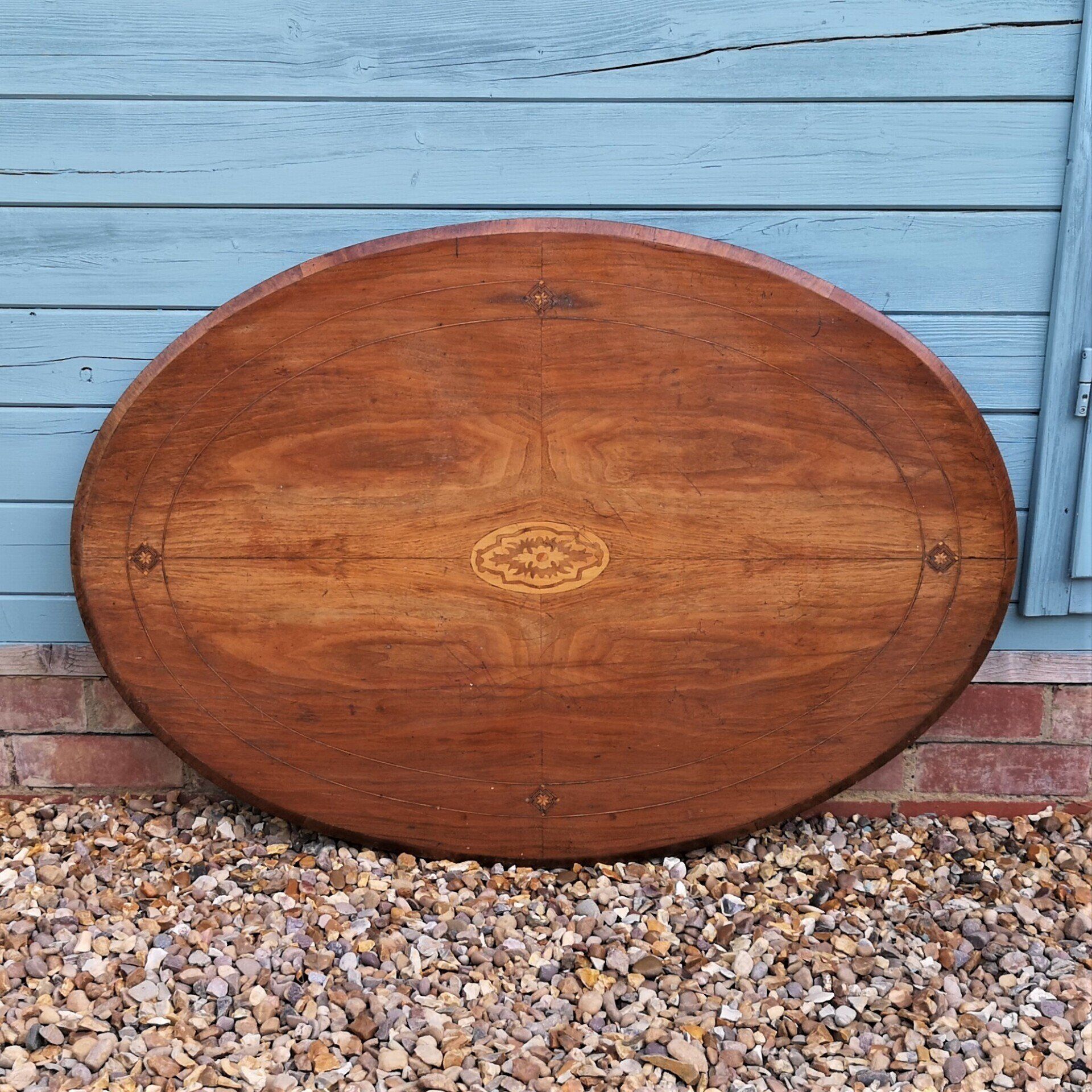 An oval wooden table is sitting on a pile of gravel in front of a blue building.