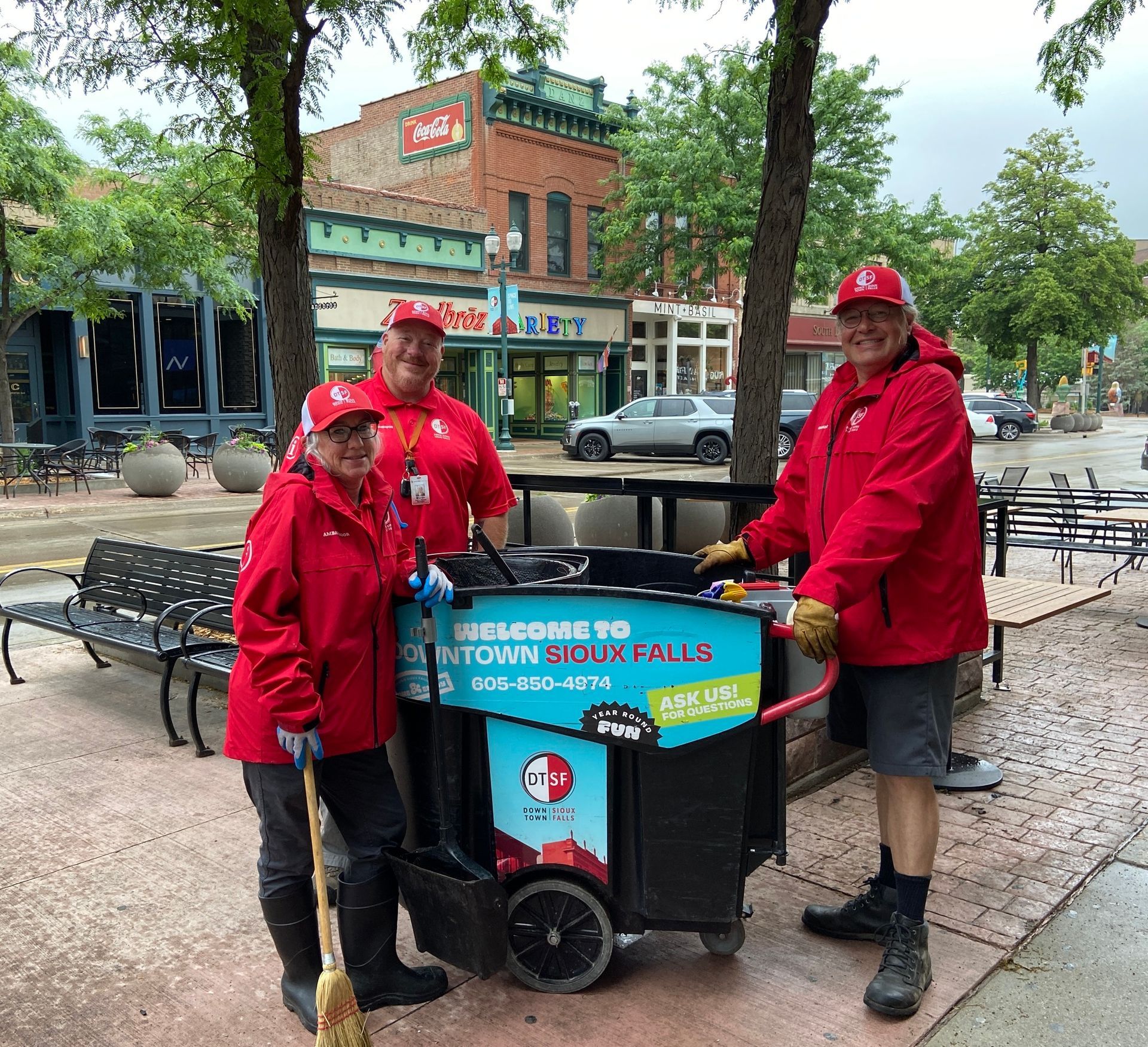Three people in red jackets with a cleaning cart on a city street.