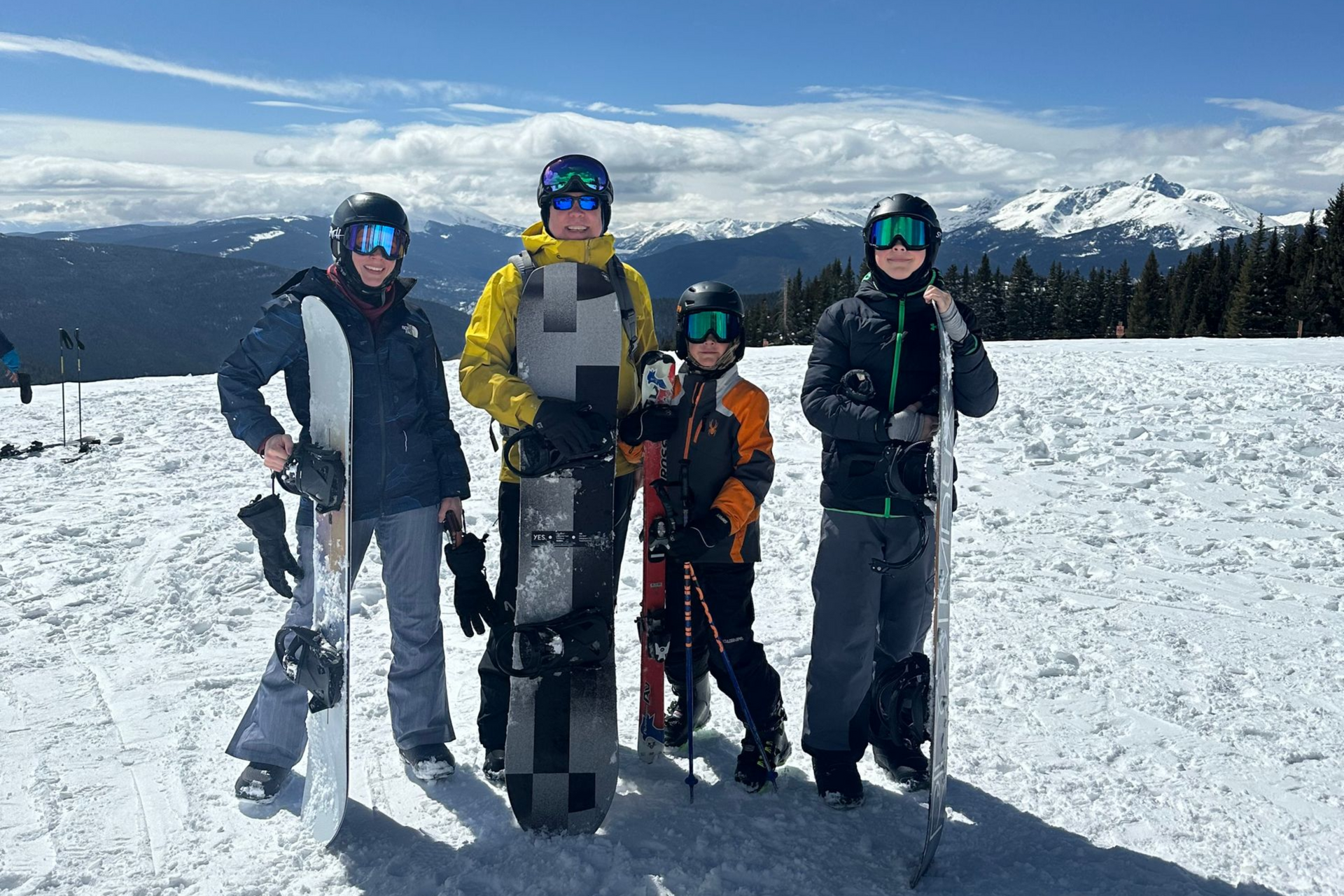 Group of people on a snowy mountain slope, wearing ski gear, posing with snowboards and skis. Blue sky and mountains in background.
