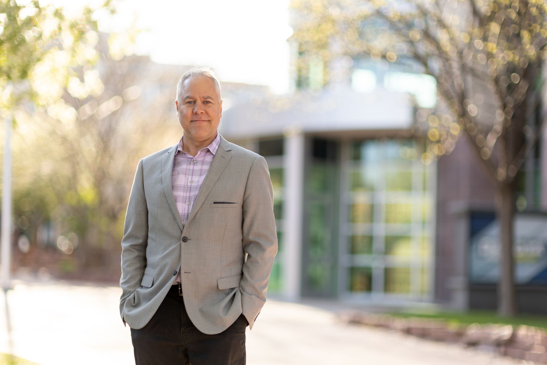 Joe in gray suit standing outside, hands in pockets, looking down. Building in background.