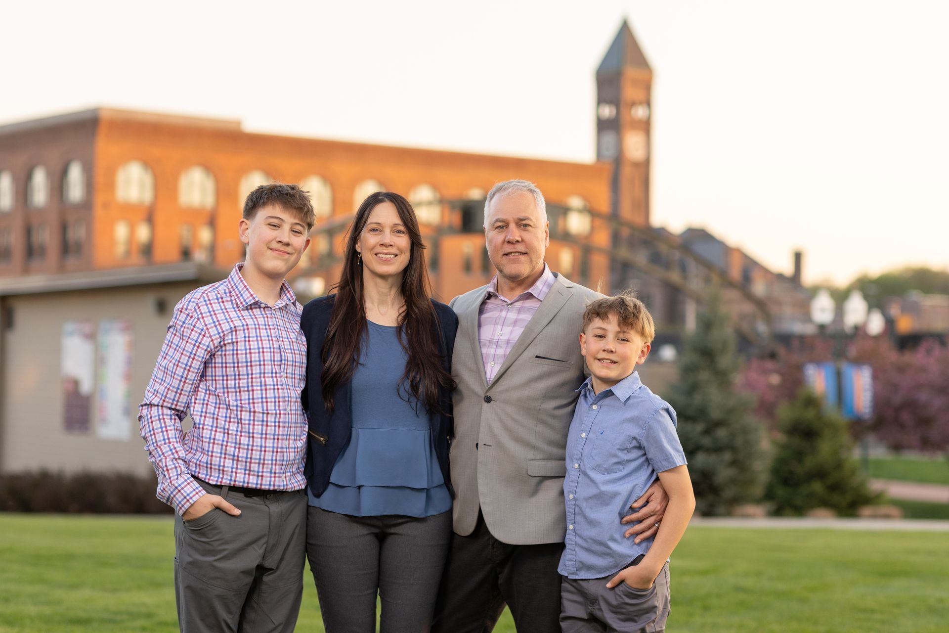 The Batcheller family of four smiles in front of a brick building with clock tower in a park setting at dusk.