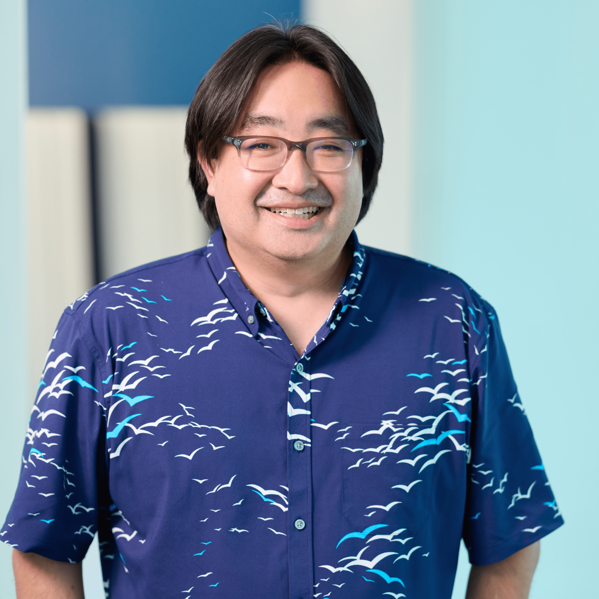 Lead Principal of Hawaii accounting firm Denny Nakayama stands in front of a light blue background wearing a dark blue shirt with bright blue and white birds.