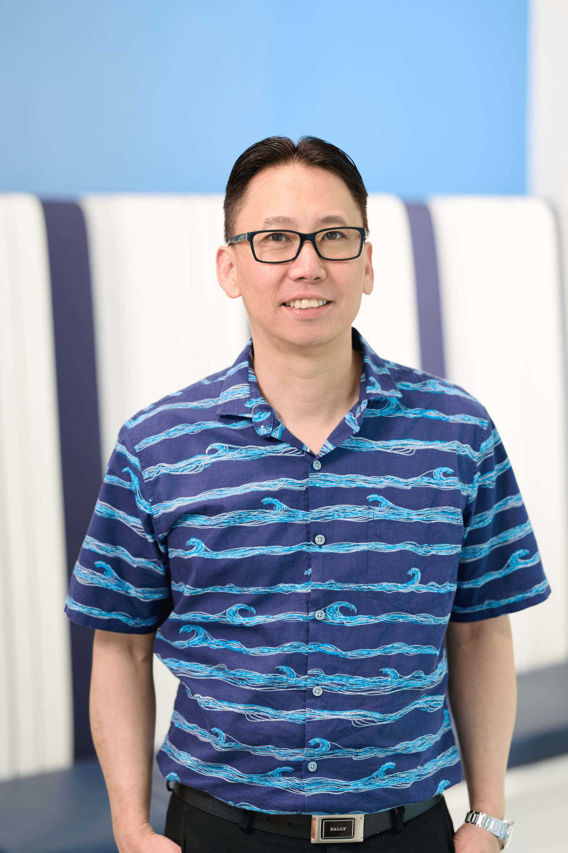 Managing Partner at N&K CPA accounting firm Blake Isobe stands in the office wearing a dark blue hawaiian shirt with a lighter blue wave pattern wearing black glasses and smiling at the camera.