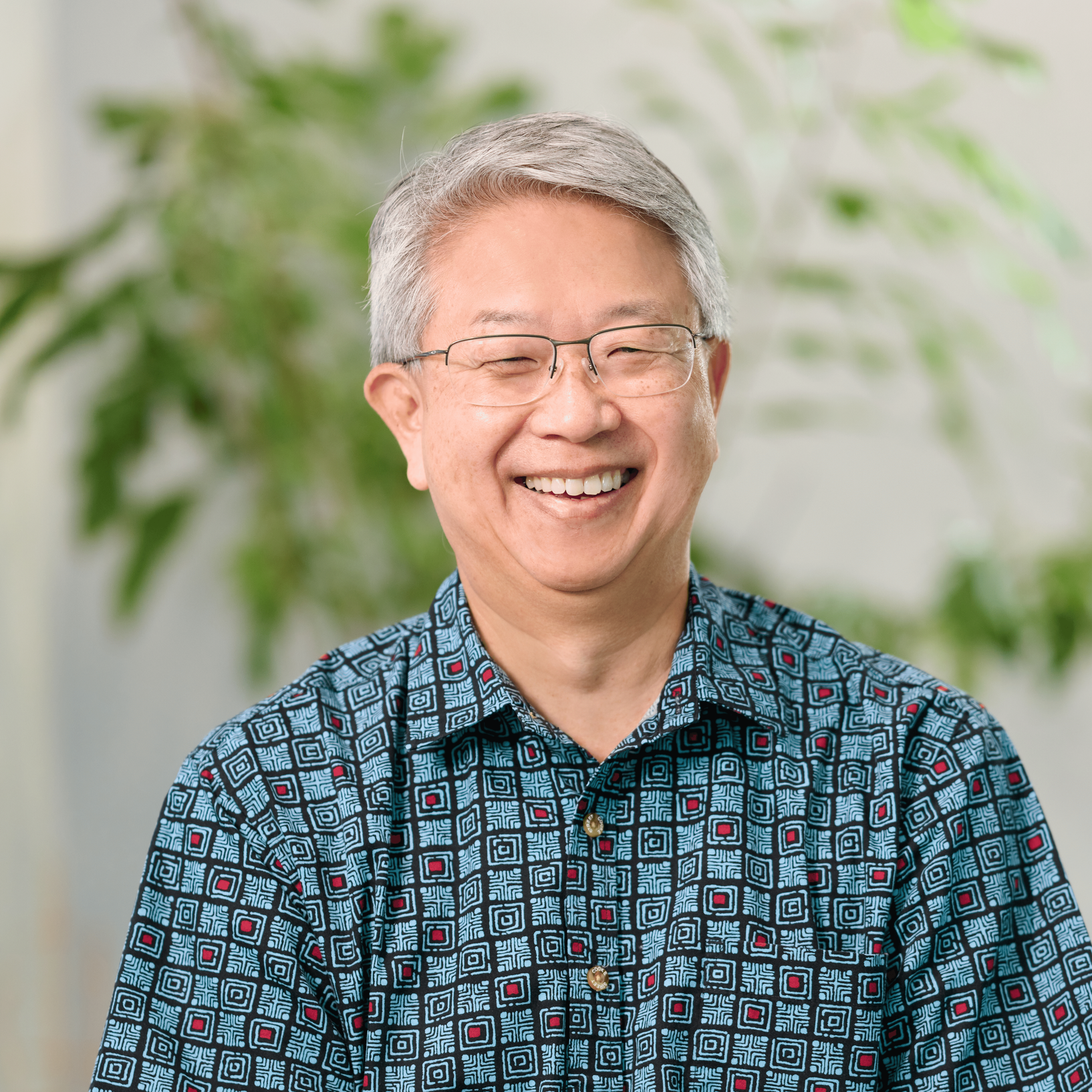 Lead CPA and accounting firm Michael Tanaka wearing a patterned blue, black, and red aloha shirt standing in front of an indoor tree .