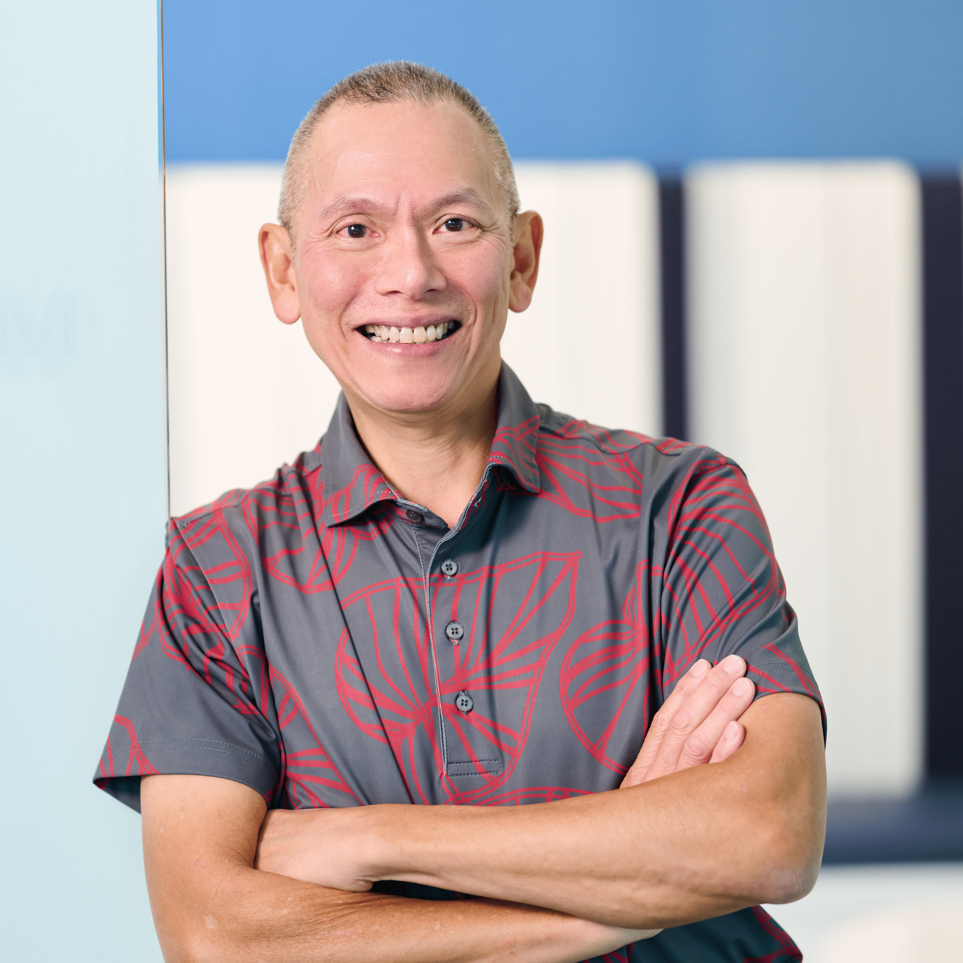 Lead accounting principal Lawrence Chew wearing a gray aloha shirt with a red monstera leaf outline, leaning against a light blue wall.