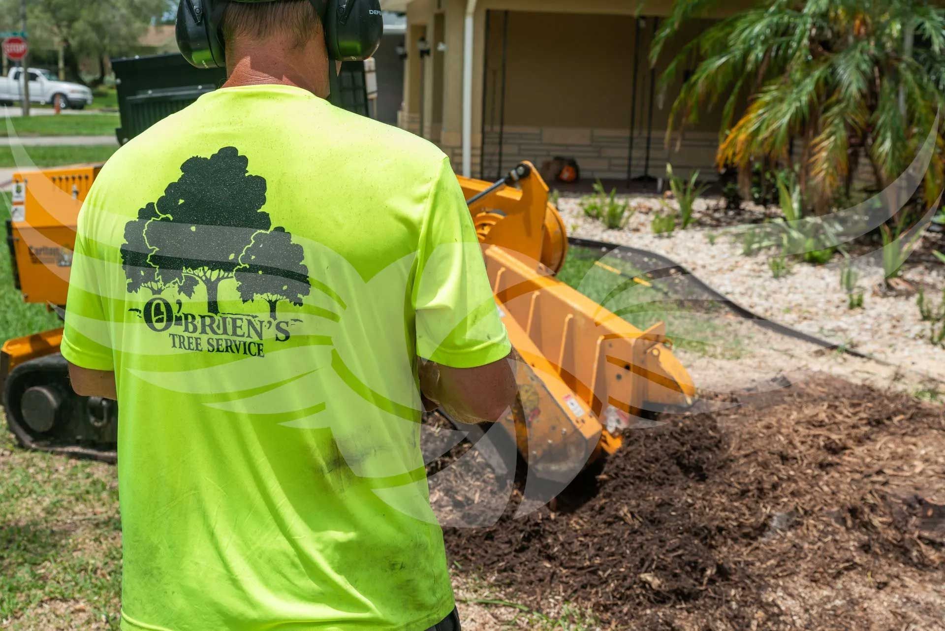 A man in a neon yellow shirt is standing next to a tree stump grinder.