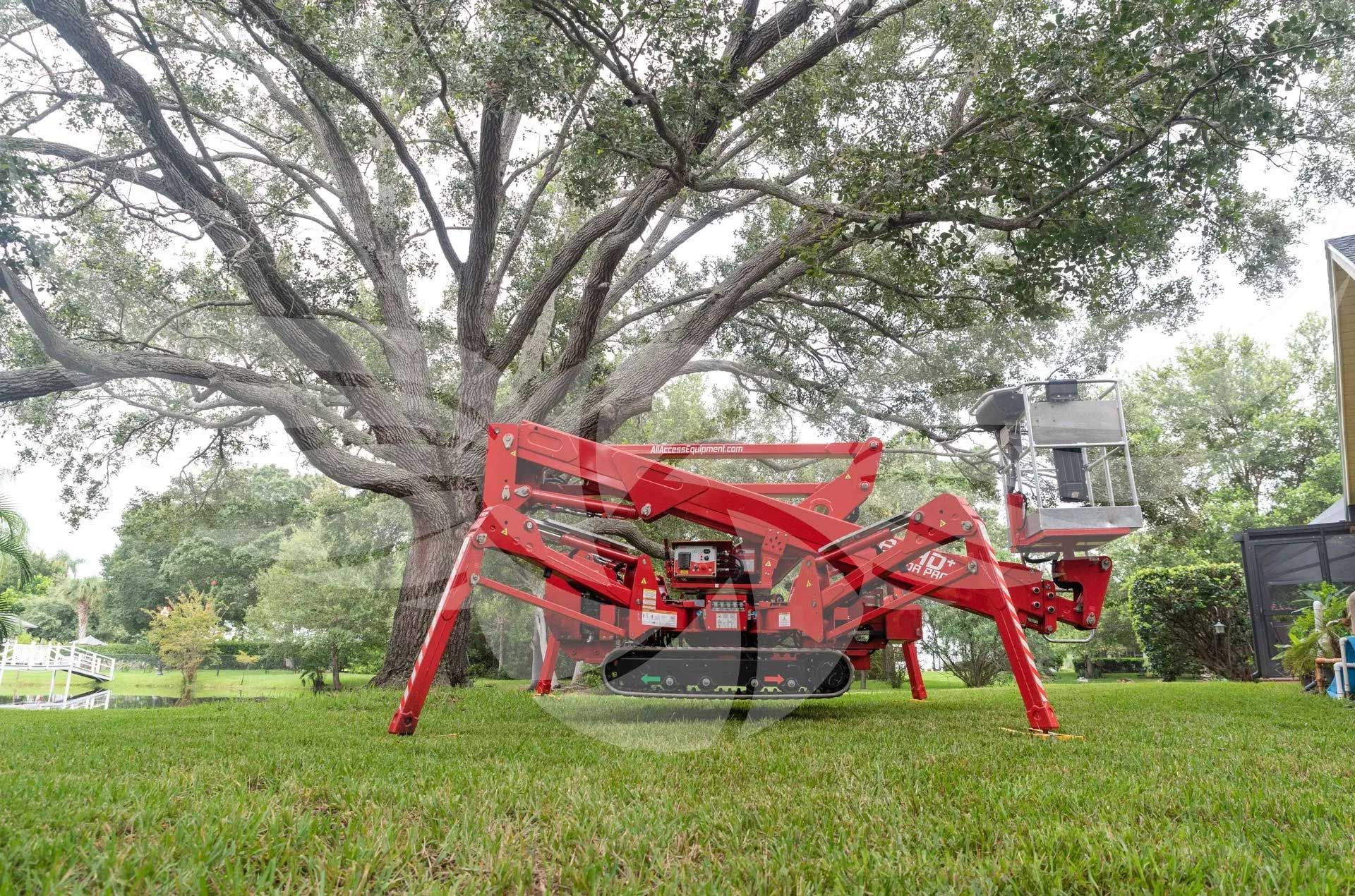 A red spider lift is sitting in the grass in front of a tree.