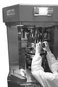 Person in lab coat adjusts machinery, likely a pill press, in a pharmaceutical setting.