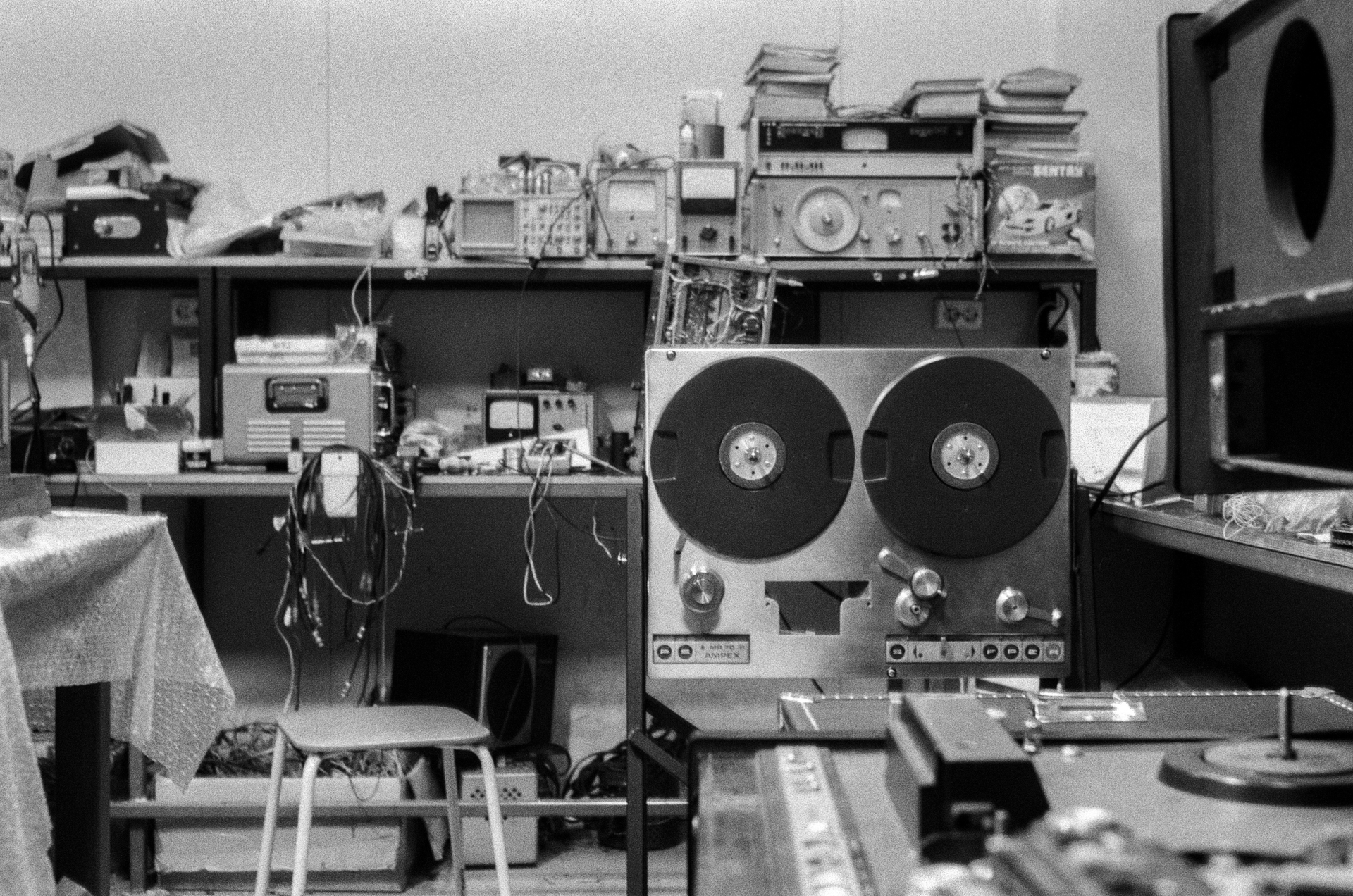 Black and white photo of a cluttered electronics workshop with a reel-to-reel tape recorder as a central focus.