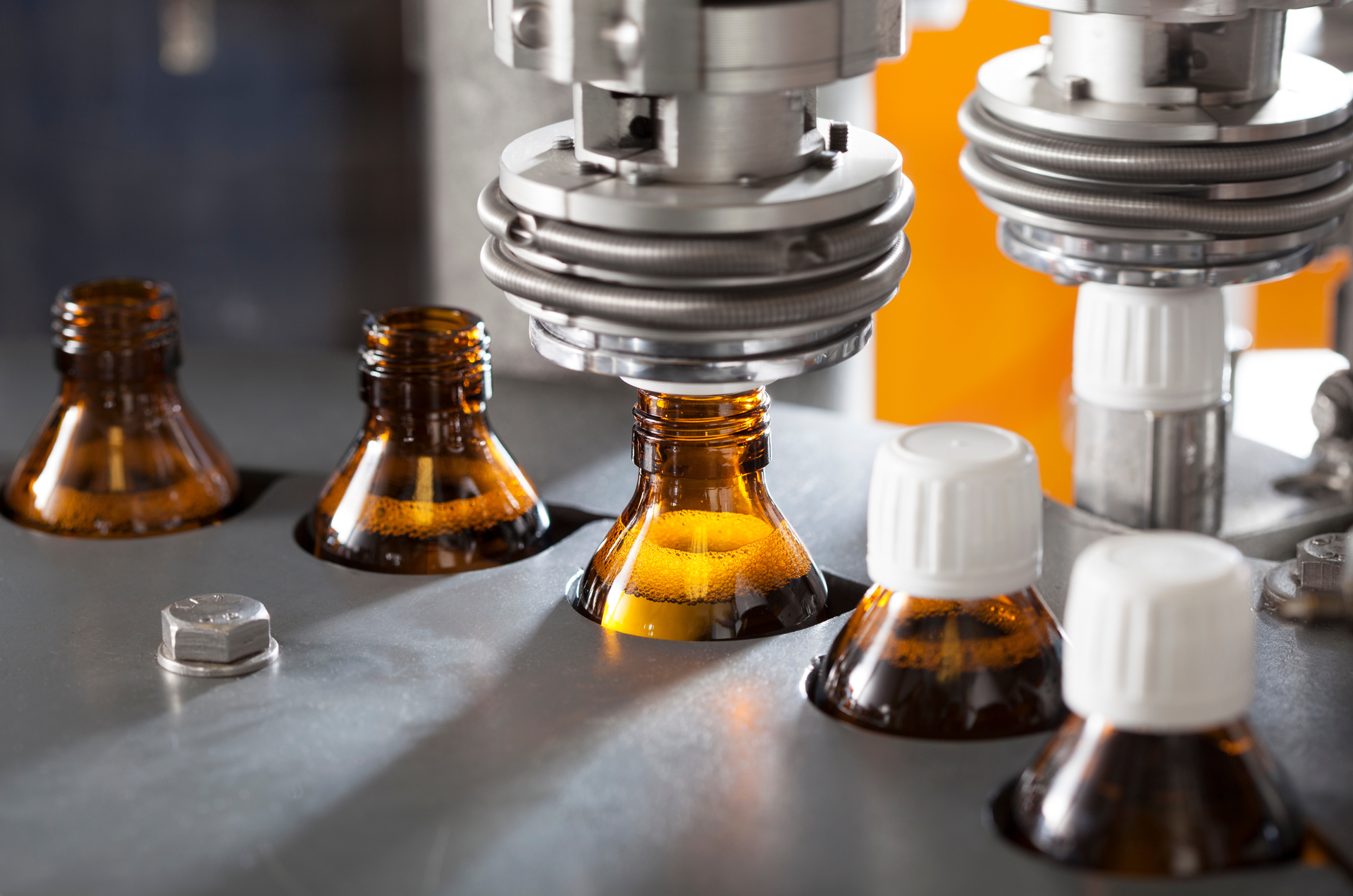 Automated bottle-capping machine in operation. Brown glass bottles are filled and sealed with white caps on a metal surface.