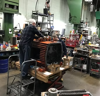 Two mechanics working on a large engine block in a workshop, surrounded by tools and equipment.