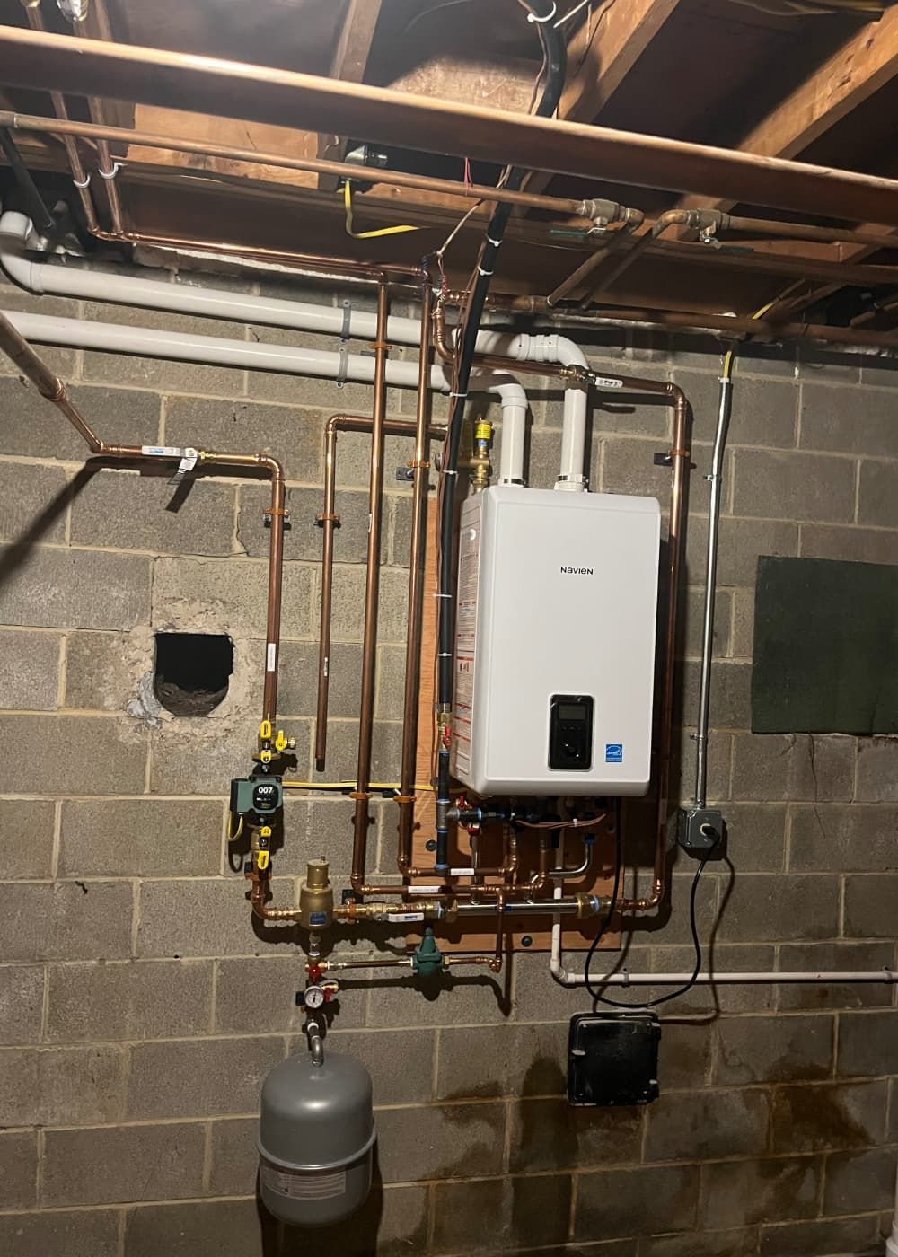 A white water heater is mounted to a brick wall in a basement.