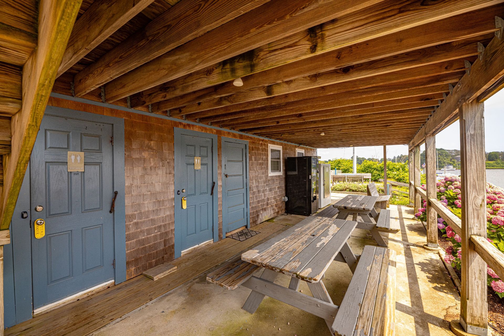 A wooden porch with a picnic table and benches under a wooden roof.