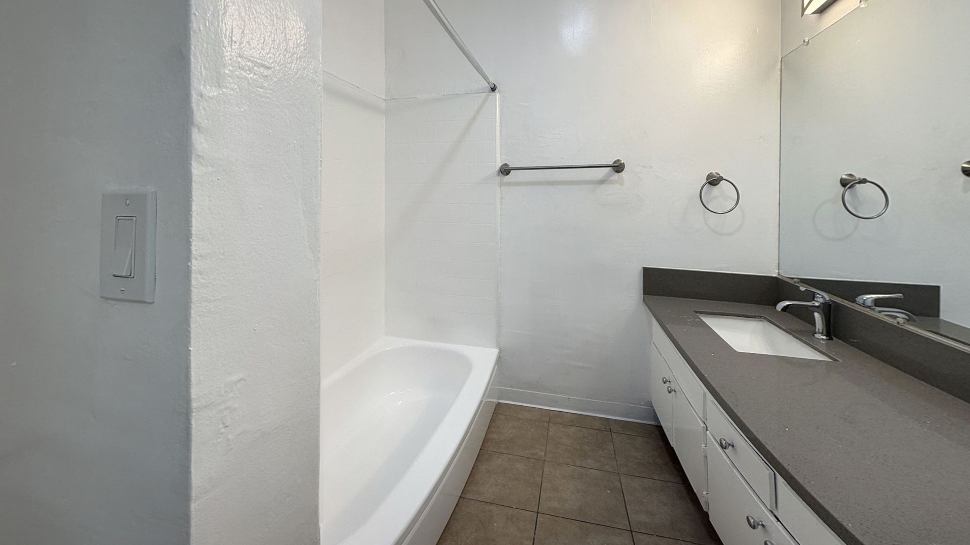 Bathroom with a white bathtub, vanity, and mirror. Gray countertop, silver towel rack and ring holders, white walls.