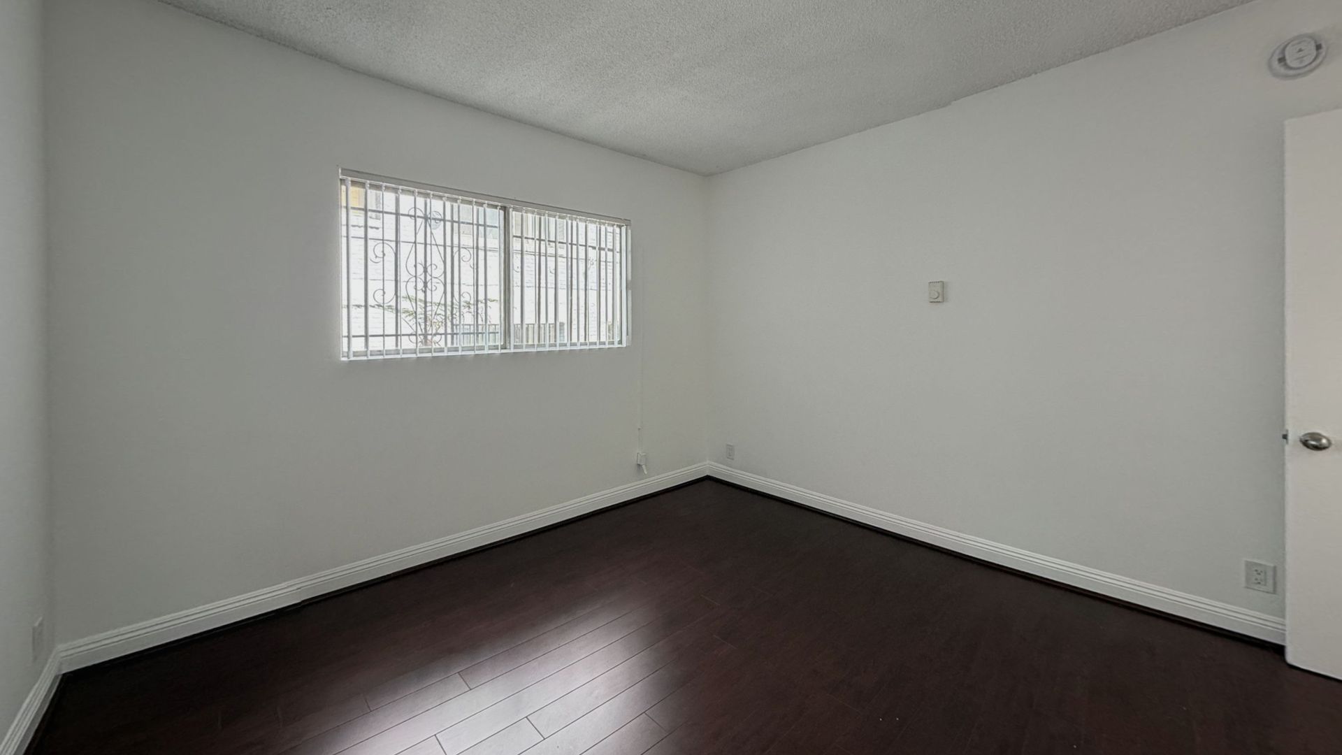 Empty room with dark wood floor, white walls, window with security bars, and a closed door.