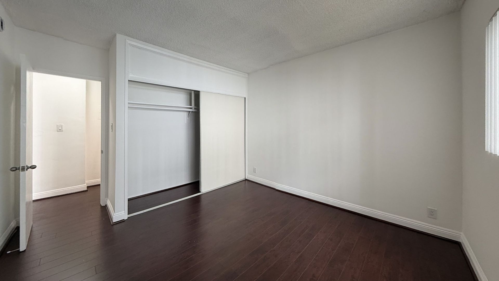 Empty bedroom with dark hardwood floors, white walls, closet, and doorway.