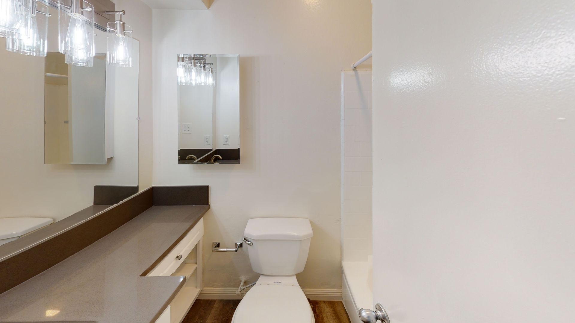 Bathroom with a toilet, sink, mirror, and shower, with white walls and a wood-look floor.