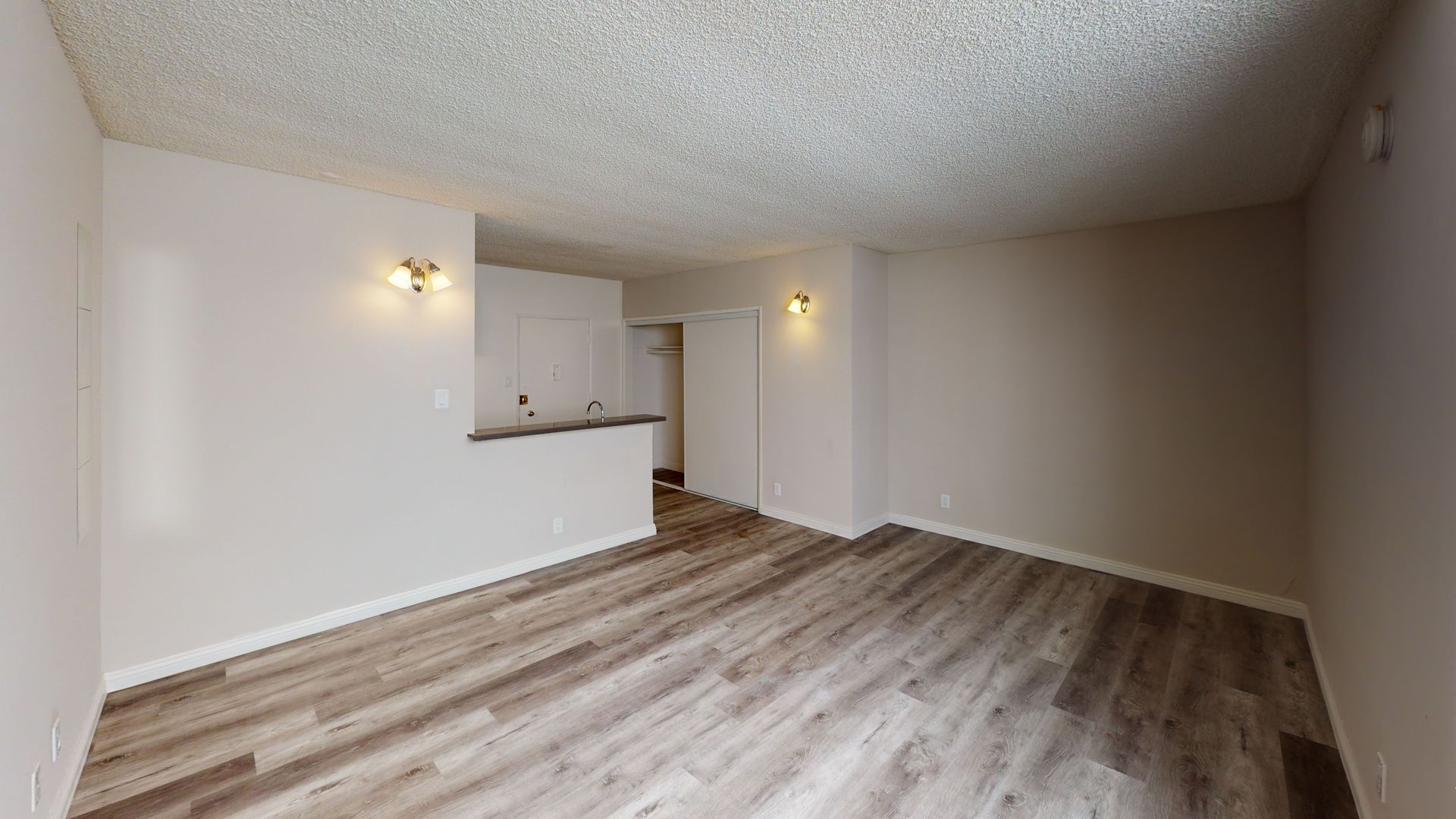 Empty, neutral-toned living space with wood-look flooring and a pass-through to a kitchen area.