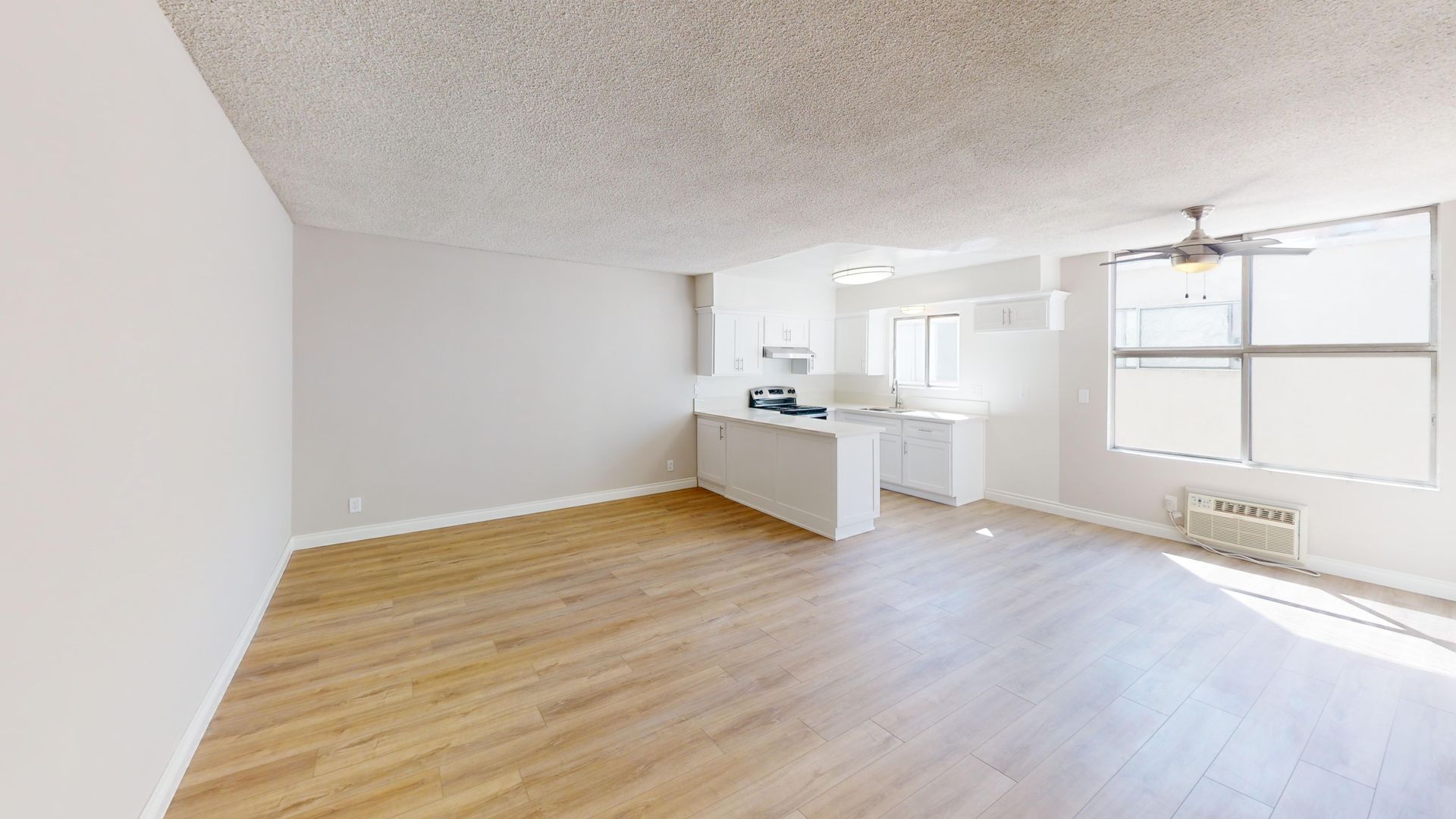 Empty, sunlit apartment interior with light wood floors, a small kitchen, and a large window.