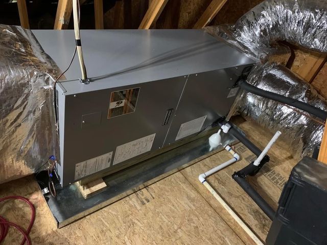 A gray HVAC air handler unit in an attic, connected to silver insulated ducts and white PVC drain pipes.