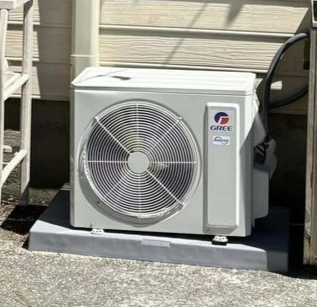 A white Gree brand outdoor air conditioner unit sits on a grey plastic pad against a beige house wall.