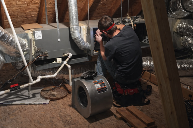 A technician kneels in an attic to repair an HVAC unit, surrounded by silver ducts and piping equipment.