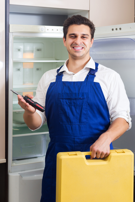 A man is standing in front of an empty refrigerator holding a screwdriver and a toolbox.
