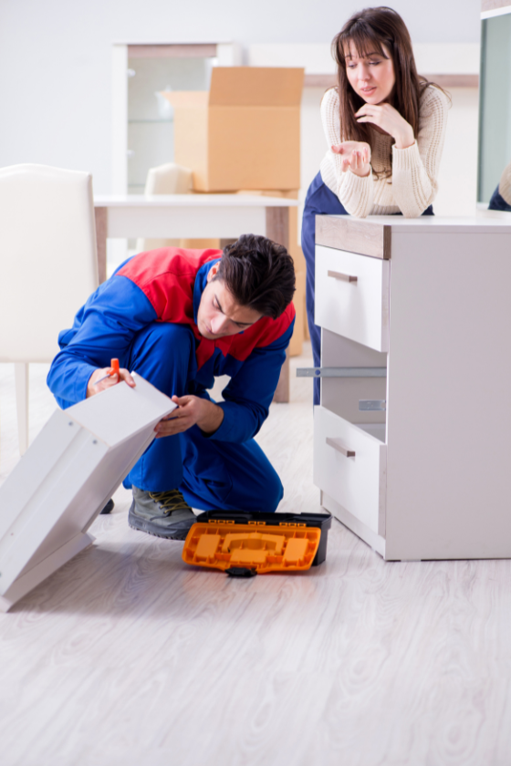 A man is fixing a piece of furniture in a living room while a woman watches.