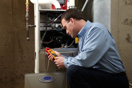 A man is kneeling down in front of a heater while holding a multimeter.