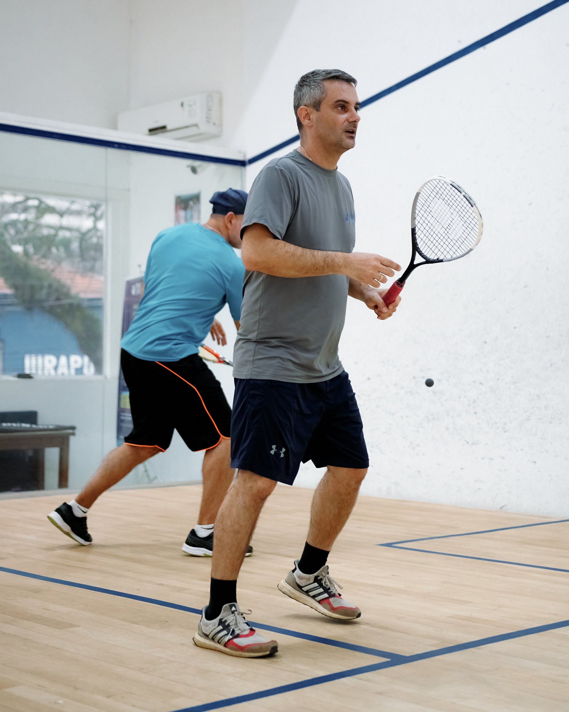 Um homem de camisa cinza se prepara para bater na bola de squash, enquanto o outro de camisa azul corre para os fundos.
