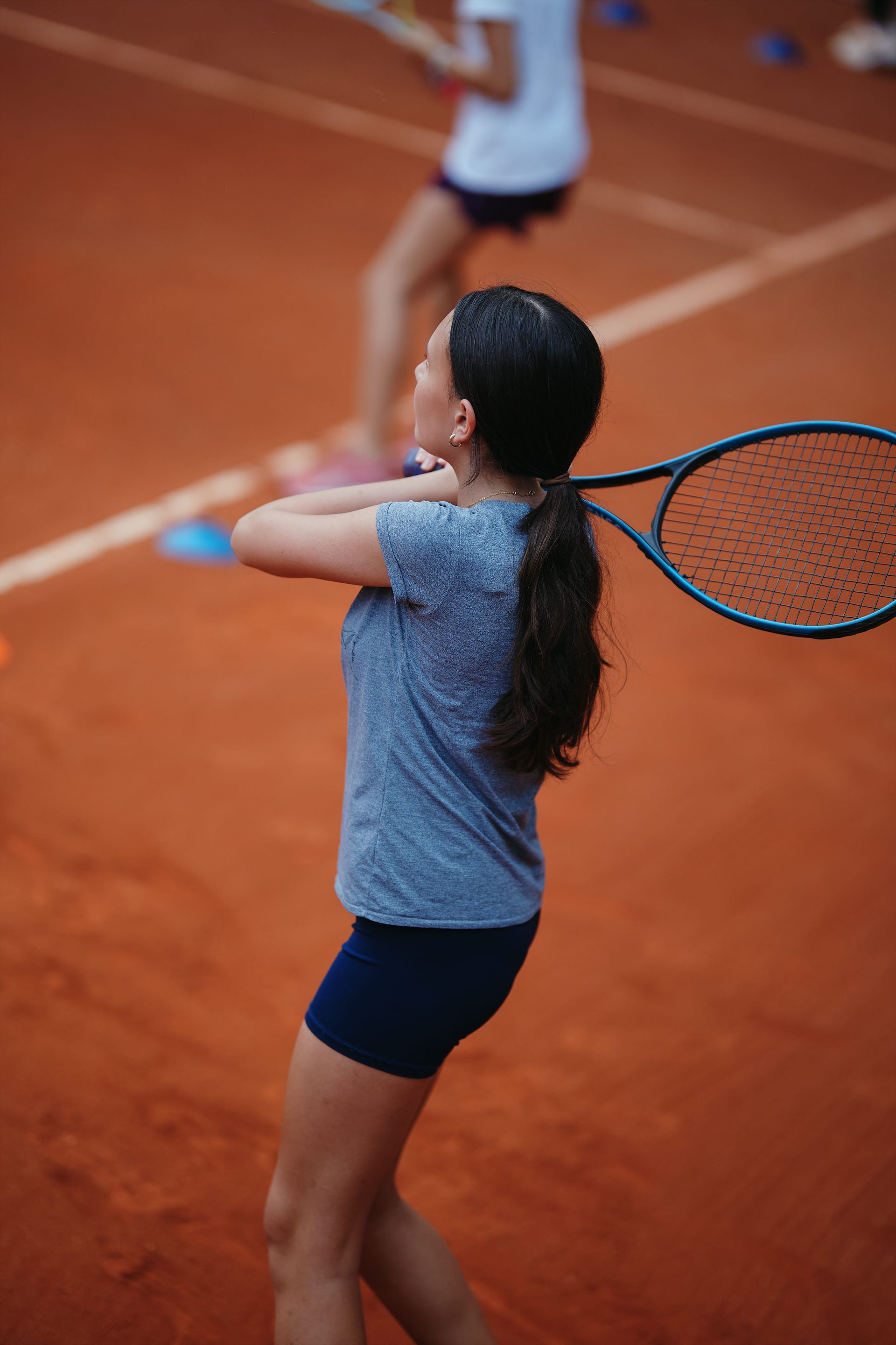 Menina de camisa azul e shorts em uma quadra de tênis de saibro, balançando uma raquete. Outra pessoa desfocada ao fundo.