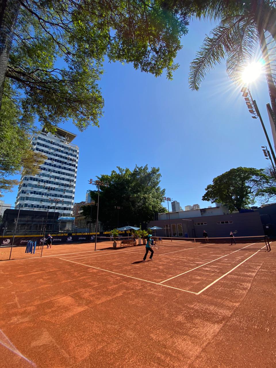 Jogador de tênis em uma quadra de saibro sob um sol brilhante. Um prédio alto é visível ao fundo, emoldurado por árvores.