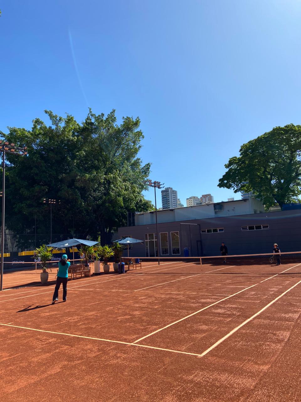 Uma pessoa jogando tênis em uma quadra de saibro em um dia ensolarado. Árvores e prédios ao fundo.