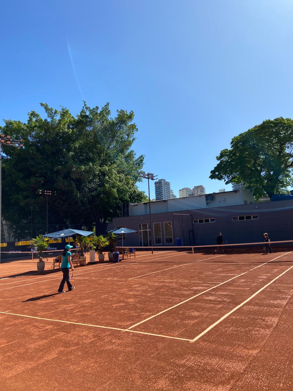 Uma pessoa joga tênis em uma quadra de saibro em um dia ensolarado. Ao fundo, árvores e um prédio.
