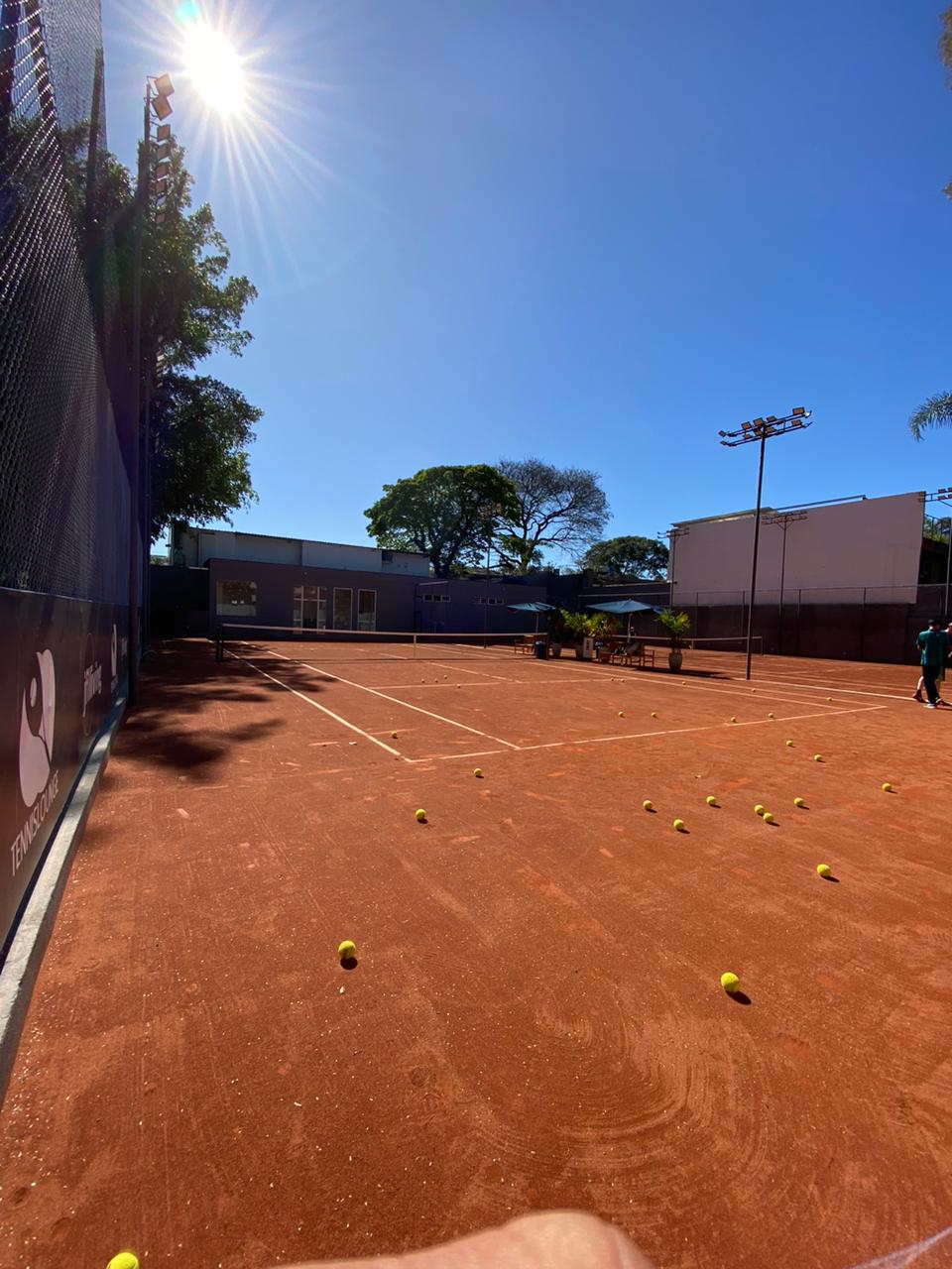 Uma quadra de tênis de saibro banhada pela luz do sol com bolas de tênis espalhadas; uma cerca e um prédio ao fundo.