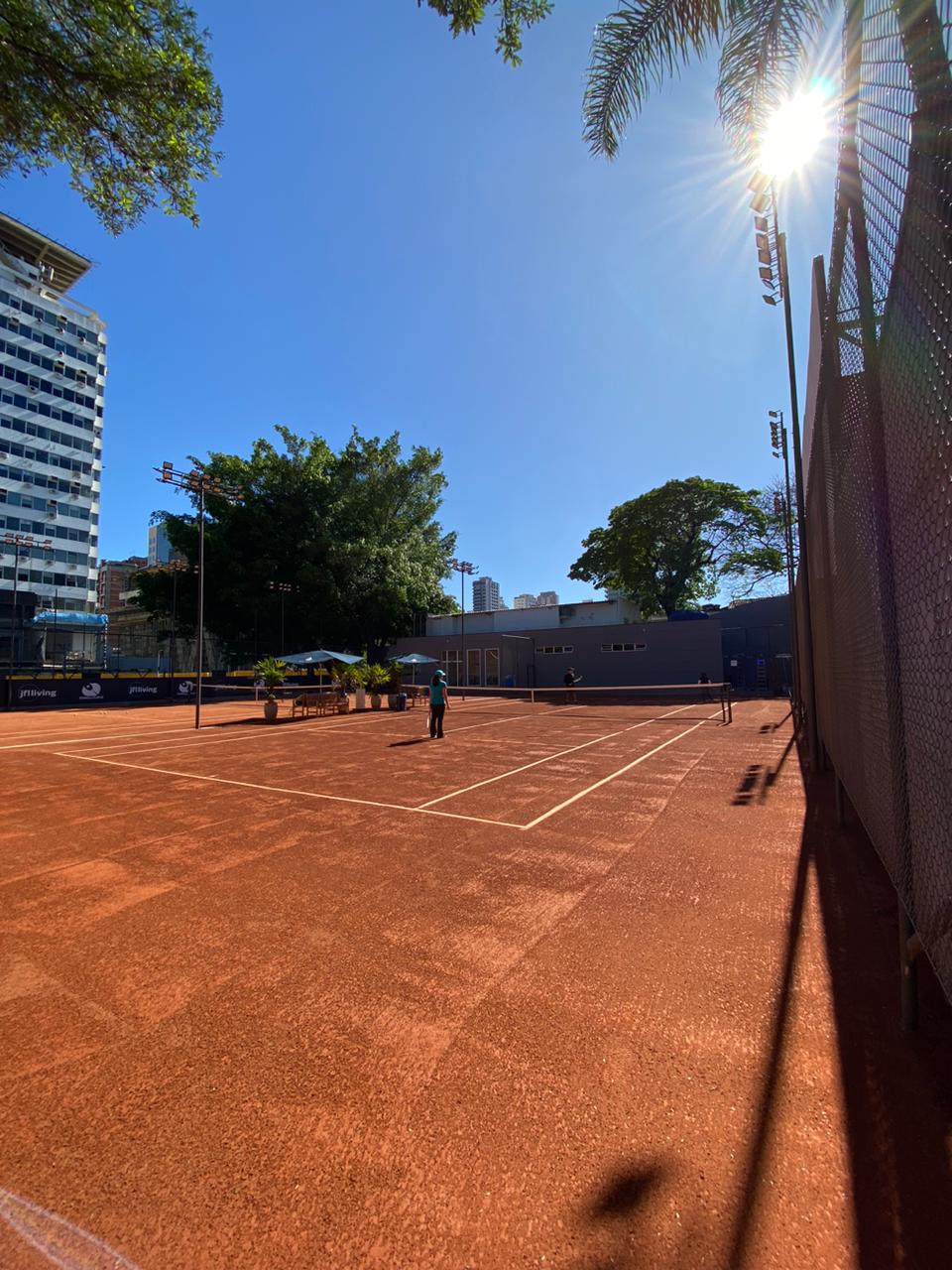 Quadra de tênis de saibro em um dia ensolarado, cercada por uma cerca de arame, árvores e prédios.