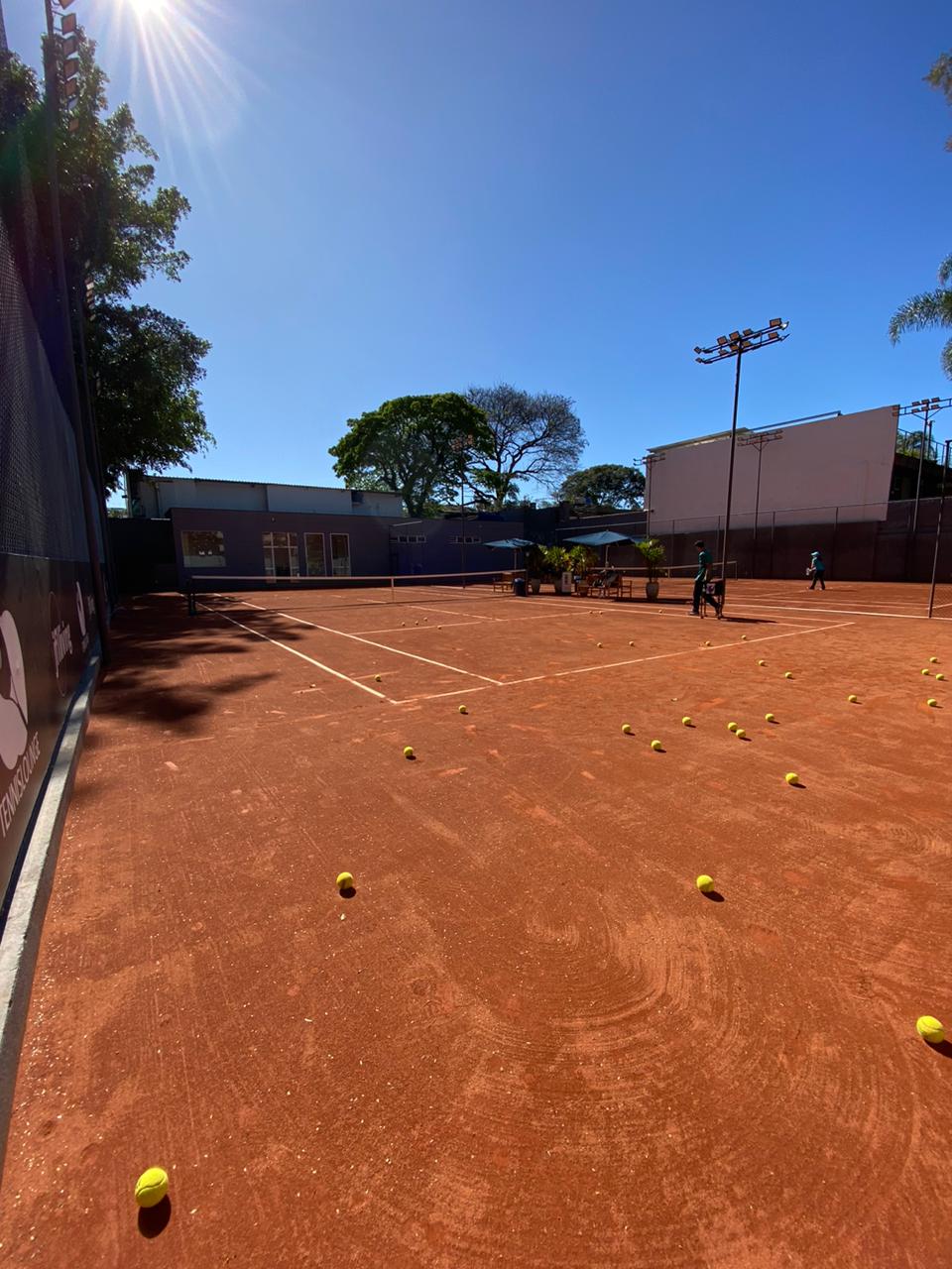 Quadra de tênis com bolas amarelas espalhadas sobre saibro vermelho sob um céu azul brilhante. Jogadores ao fundo.