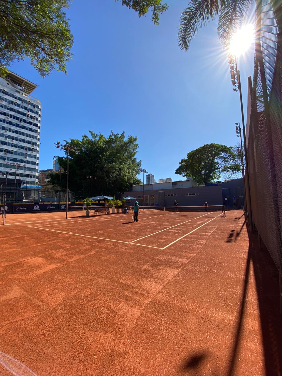 Quadra de tênis de saibro vermelho em um dia ensolarado, jogadores visíveis e prédios altos ao fundo.