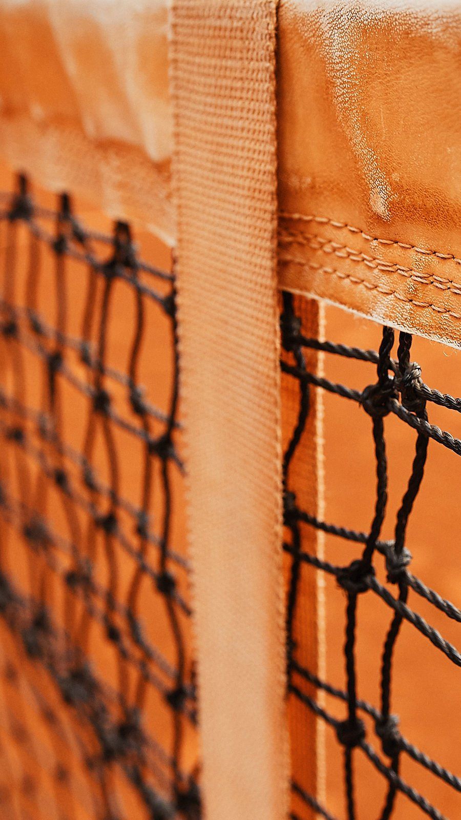 Close de uma rede de tênis com suporte laranja e tela preta. A rede está posicionada contra o fundo de uma quadra de saibro.