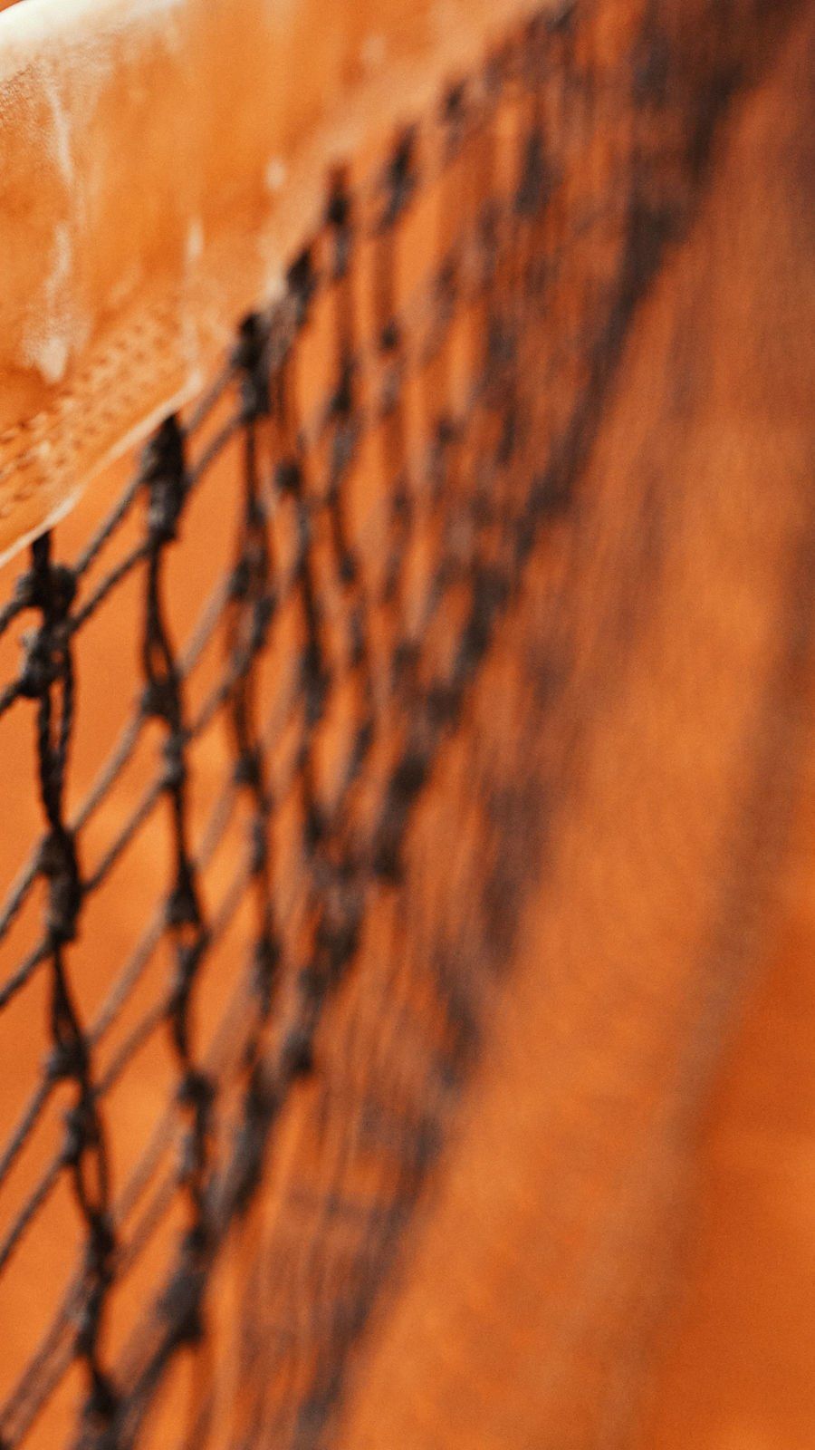 Rede de tênis e quadra de saibro ao fundo, close-up. Rede preta contra uma superfície de saibro laranja desfocada.